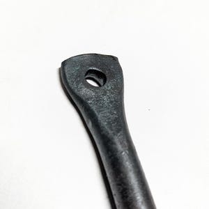 Könnte beinhalten: Nahaufnahme eines dunkelgrauen Metallgriffs mit einem Loch oben. Der Griff hat eine leicht strukturierte Oberfläche und eine abgerundete Form, was darauf hindeutet, dass es sich um ein Werkzeug oder Teil eines größeren Mechanismus handelt.