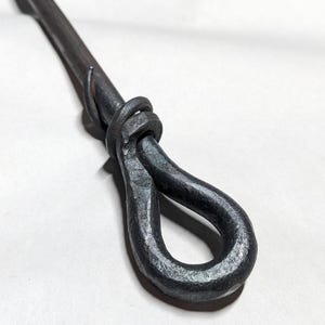 Könnte beinhalten: Handgeschmiedeter Metall-Kaminhaken mit Schlaufengriff. Der dunkelgraue Stahl hat ein strukturiertes, gehämmertes Aussehen. Der Griff ist zu einer Schlaufe geformt, und der Schaft ist eine lange, schlanke Stange.