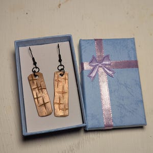 May include: Copper-colored rectangular earrings with a cross-hatch design are displayed in a light blue gift box. The earrings have black ear wires. A matching gift box with a lavender ribbon and bow is next to the open box.