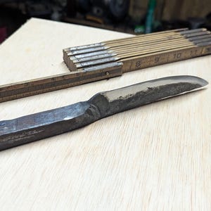 May include: A hand-forged, dark metal knife with a curved blade rests on a light wood surface, alongside a vintage wooden folding ruler. The ruler is partially open, displaying centimetre markings. The knife's handle is thick and sturdy.