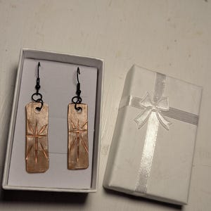 May include: A pair of copper-colored rectangular earrings with a starburst design, displayed in a white gift box. The earrings have black hooks and are next to a closed white gift box with a silver ribbon.
