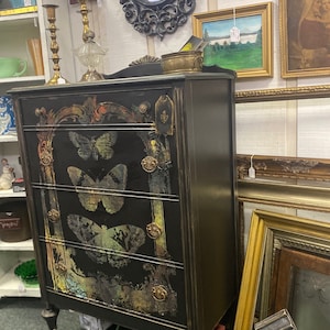 May include: A black dresser with four drawers decorated with gold accents and butterfly designs. The dresser has a dark wood finish and sits on four legs. The dresser is in a shop with other furniture and decor.