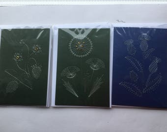 May include: Three greeting cards with different designs. The first card is green with a white embroidered dandelion design. The second card is green with a white embroidered dandelion design. The third card is blue with a white embroidered thistle design.