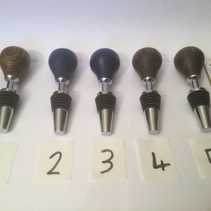 May include: Five wooden wine bottle stoppers with silver bases and black rubber seals. The wooden tops are in shades of brown and blue. Each stopper is numbered on a small white square.