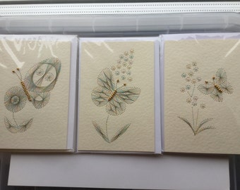 May include: Three handmade greeting cards featuring string art designs of butterflies and flowers. The cards are on a cream-colored paper, with colorful thread used to create the designs. Each card is individually packaged in a clear plastic sleeve.