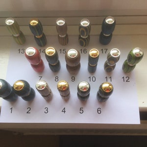 Op de afbeelding: Een set van 17 houten flessenstoppers met zilveren en gouden accenten. De stoppers zijn gerangschikt in twee rijen op een wit oppervlak met de nummers 1-17 eronder gedrukt.