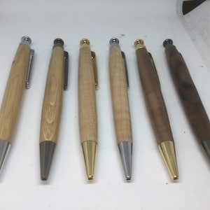 Puede incluir: Siete bolígrafos de madera con detalles metálicos de diferentes colores. Los bolígrafos son todos del mismo tamaño y forma, y están dispuestos en fila sobre una superficie blanca.