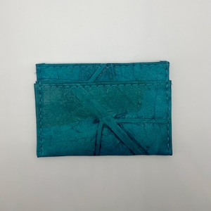 May include: A teal blue leather wallet with a textured surface. The wallet has a fold-over design and a stitched edge.