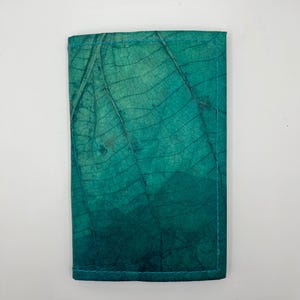 Puede incluir: Una cartera azul turquesa hecha de papel reciclado con un patrón de hojas. La cartera tiene un borde cosido y está cerrada.