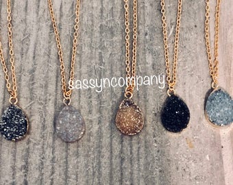 May include: Five gold chain necklaces with teardrop-shaped pendants. The pendants are made of a sparkly, speckled material in five different colors: black, gray, white, light brown, and dark gray.