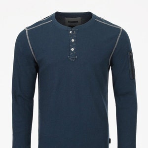 May include: A dark blue long-sleeved shirt with a waffle knit texture. The shirt has a button placket and a zippered pocket on the left sleeve. The shirt has white stitching around the neckline, cuffs, and hem.