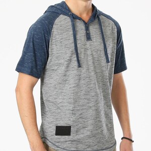 May include: A gray and blue short-sleeve hooded shirt. The shirt has a gray body with blue sleeves and a matching hood. It features a button-up neckline and a small black patch near the hem. The shirt is made of a soft, textured fabric.