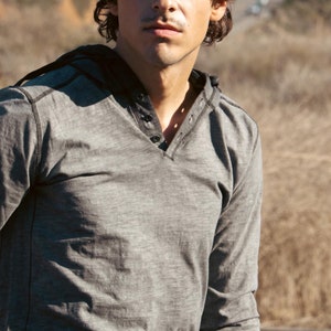 May include: A man wearing a gray long-sleeved shirt with a v-neck and a button closure. The shirt has a textured look and is slightly wrinkled.