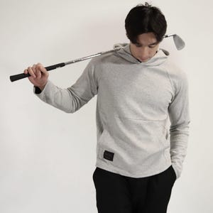 May include: A light grey long-sleeve hooded sweatshirt with a textured diamond pattern, a front kangaroo pocket, and a small black rectangular label on the lower left side. The garment features raglan sleeves and a hood with a crossover neckline.