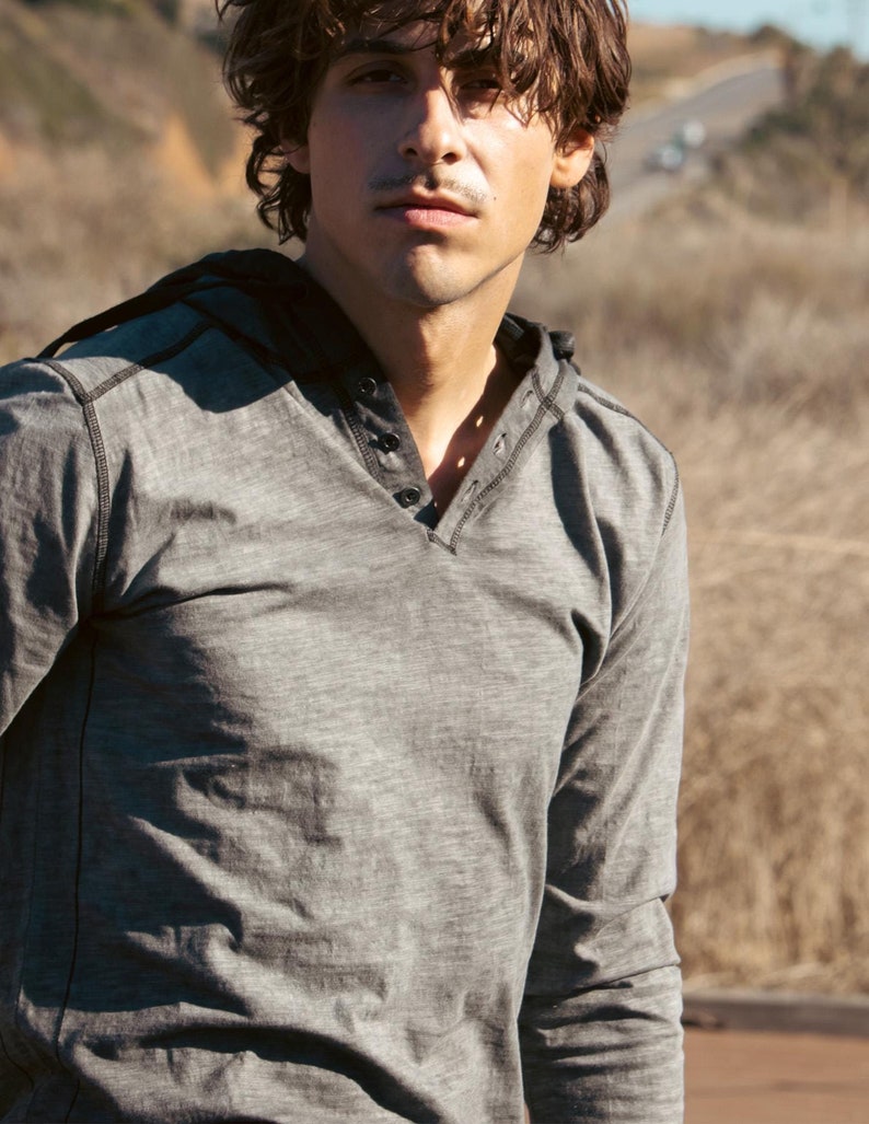 May include: A man wearing a gray long-sleeved shirt with a v-neck and a button closure. The shirt has a textured look and is slightly wrinkled.