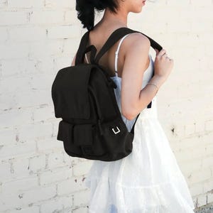 May include: A solid black backpack featuring a top flap closure, adjustable shoulder straps, and two front pockets with rectangular silver-toned metal hardware accents on the side, constructed from a durable fabric material.