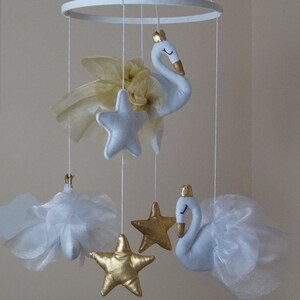 Puede incluir: M&oacute;vil de oro blanco con dos cisnes blancos, estrellas doradas y nubes blancas. El m&oacute;vil est&aacute; colgado de un anillo blanco.