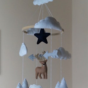 May include: A handmade baby mobile featuring felt cutouts of clouds, mountains, a deer, polar bears, a star, and arrows. The mobile is suspended from a wooden hoop with white string, creating a whimsical nursery decoration.