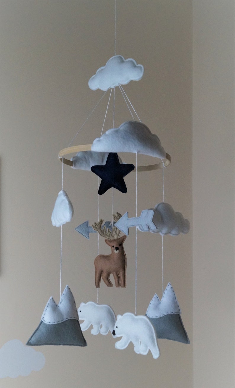 May include: A handmade baby mobile featuring felt cutouts of clouds, mountains, a deer, polar bears, a star, and arrows. The mobile is suspended from a wooden hoop with white string, creating a whimsical nursery decoration.