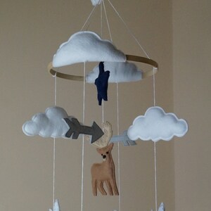 May include: A handmade baby mobile featuring felt cutouts of clouds, mountains, a deer, a polar bear, and arrows. The mobile is suspended from a wooden hoop and includes white string for hanging. The color palette includes white, brown, gray, and navy blue.