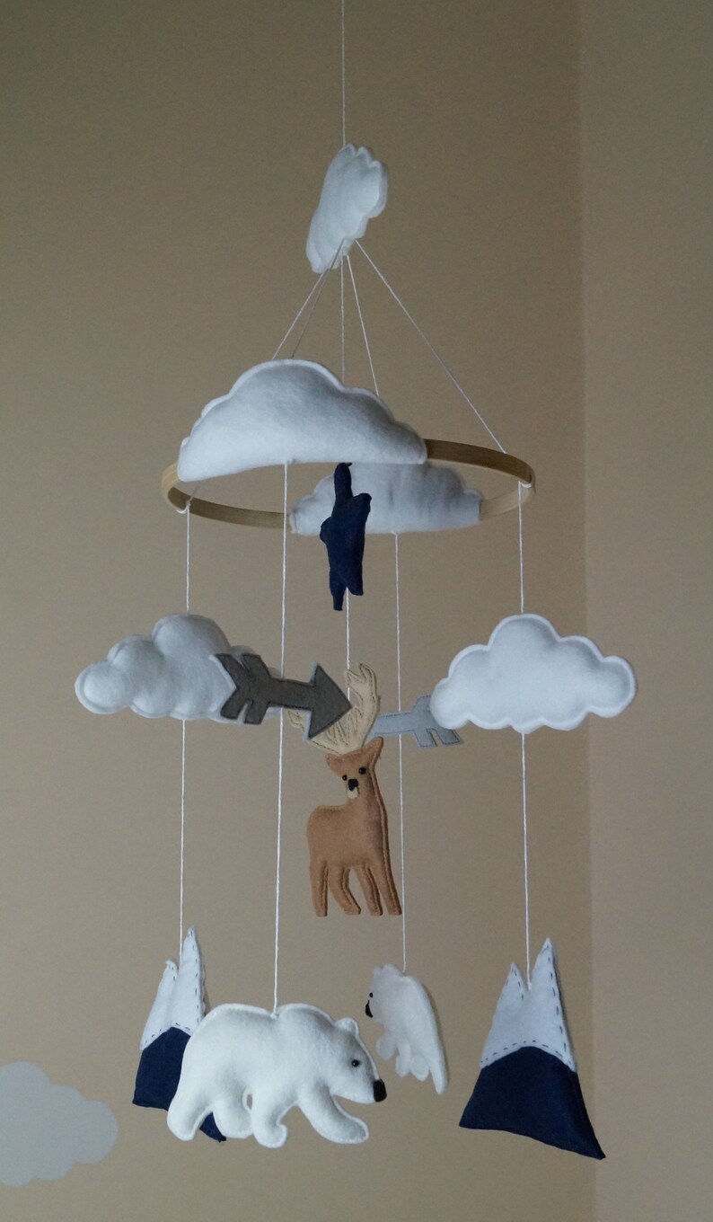 May include: A handmade baby mobile featuring felt cutouts of clouds, mountains, a deer, a polar bear, and arrows. The mobile is suspended from a wooden hoop and includes white string for hanging. The color palette includes white, brown, gray, and navy blue.