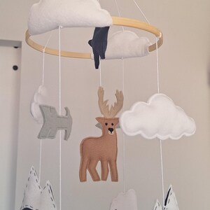 May include: A handmade baby mobile featuring felt cutouts of clouds, mountains, a deer, a polar bear, and other woodland creatures. The mobile is suspended from a wooden hoop with white string. The color palette includes white, brown, gray, and navy blue.