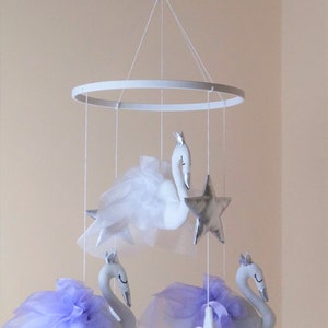 Puede incluir: Un m&oacute;vil para beb&eacute; blanco y plateado con tres cisnes blancos y tres estrellas plateadas. Los cisnes tienen tul morado y blanco unido a ellos. El m&oacute;vil est&aacute; colgado de un anillo blanco.
