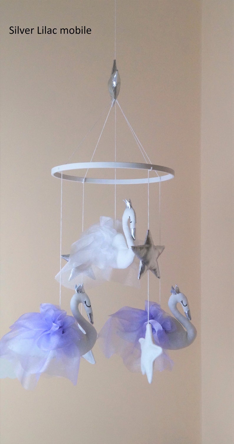 Puede incluir: Un m&oacute;vil para beb&eacute; blanco y plateado con tres cisnes blancos y tres estrellas plateadas. Los cisnes tienen tul morado y blanco unido a ellos. El m&oacute;vil est&aacute; colgado de un anillo blanco.