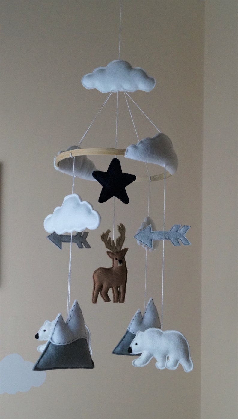 May include: A handmade baby mobile featuring felt clouds, a black star, gray arrows, a brown deer, polar bears, and gray mountains. The mobile is suspended from a wooden hoop with white string.