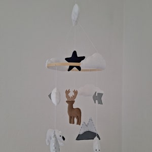 May include: A baby mobile featuring felt cutouts of a black star, white clouds, a brown moose, mountains, and polar bears. The mobile is suspended from a wooden hoop and white strings, creating a whimsical nursery decoration.
