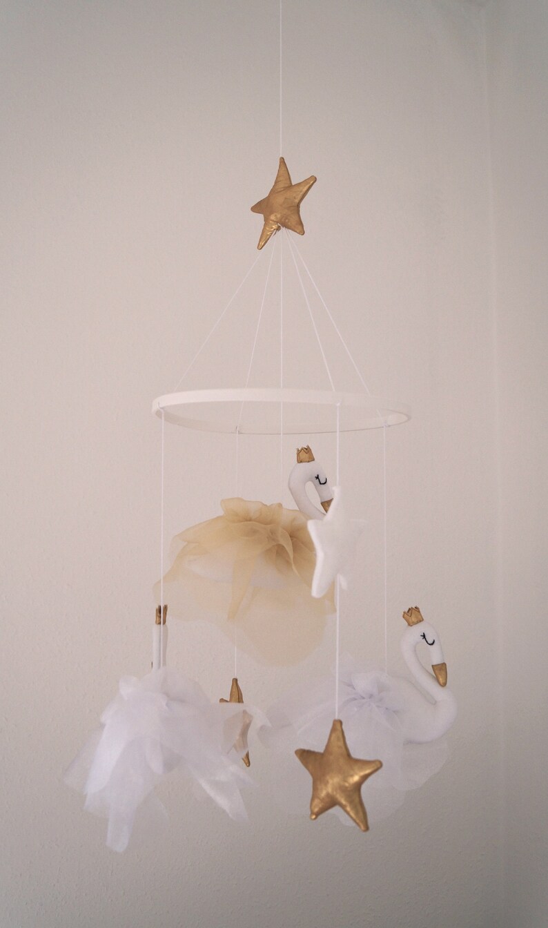 Puede incluir: Un m&oacute;vil de beb&eacute; blanco y dorado con dos cisnes blancos, tres estrellas doradas y tul blanco. El m&oacute;vil est&aacute; colgado de un techo blanco.