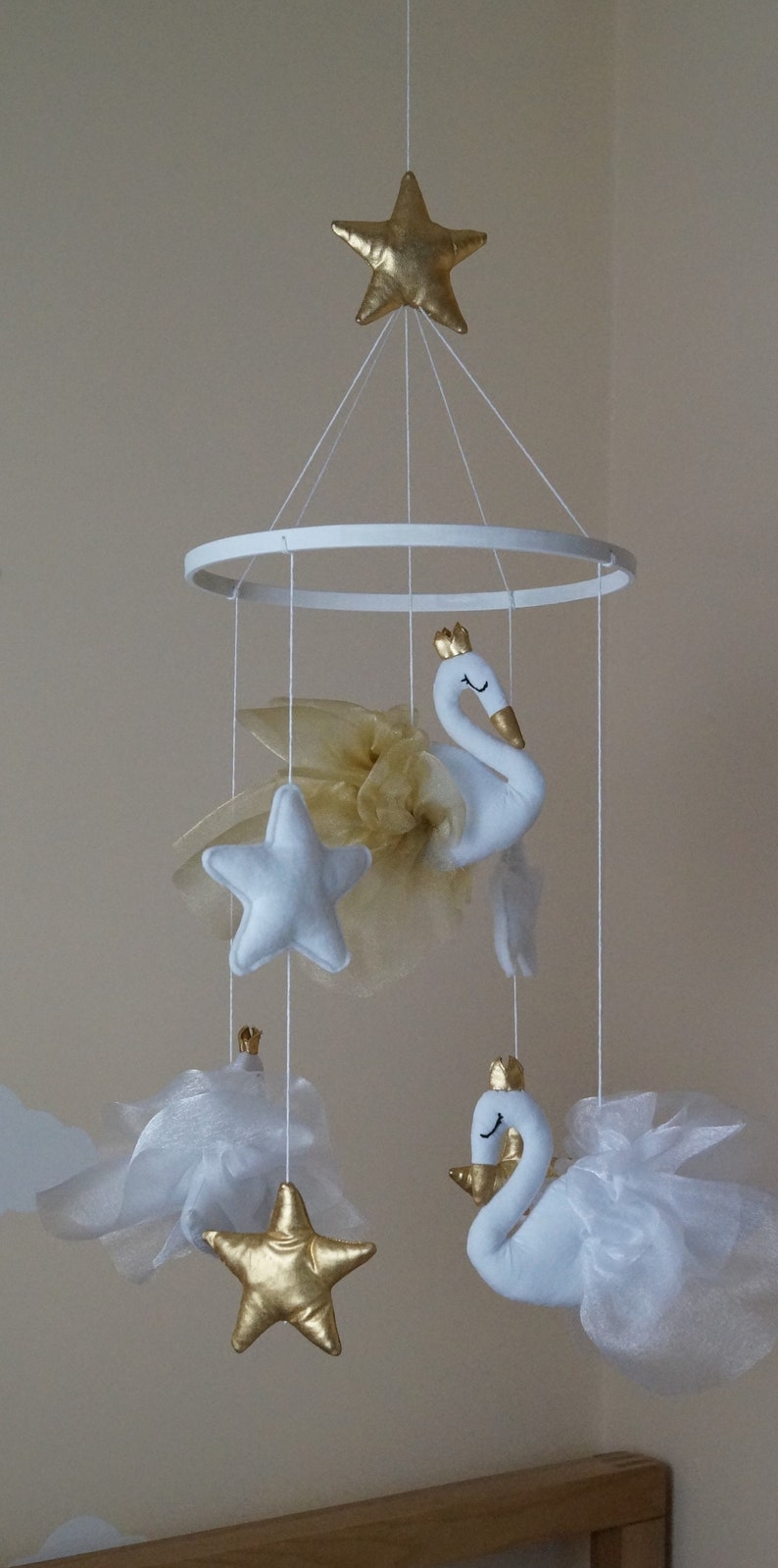 Puede incluir: Un m&oacute;vil para beb&eacute; blanco y dorado con dos cisnes blancos, tres estrellas y tul blanco. El m&oacute;vil est&aacute; colgado de un anillo blanco.