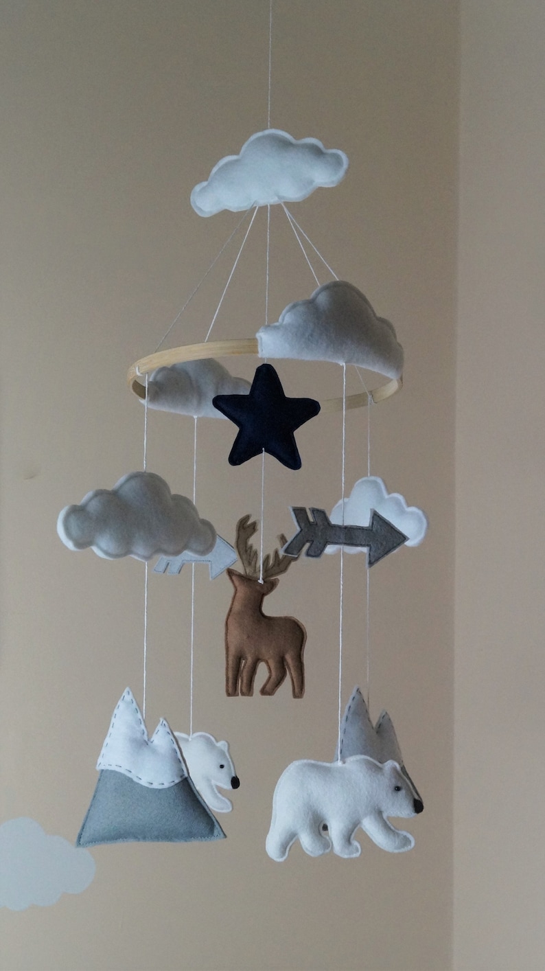 May include: A handmade baby mobile featuring felt clouds, a navy star, a brown deer, a gray arrow, mountains, and polar bears. The mobile is suspended from a wooden hoop with white strings, creating a whimsical nursery decoration.