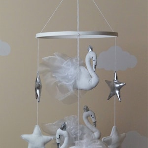 Puede incluir: M&oacute;vil de beb&eacute; blanco y plateado con tres cisnes blancos y tres estrellas plateadas. Los cisnes tienen alas de tul blanco y coronas. El m&oacute;vil est&aacute; colgado de un anillo blanco.