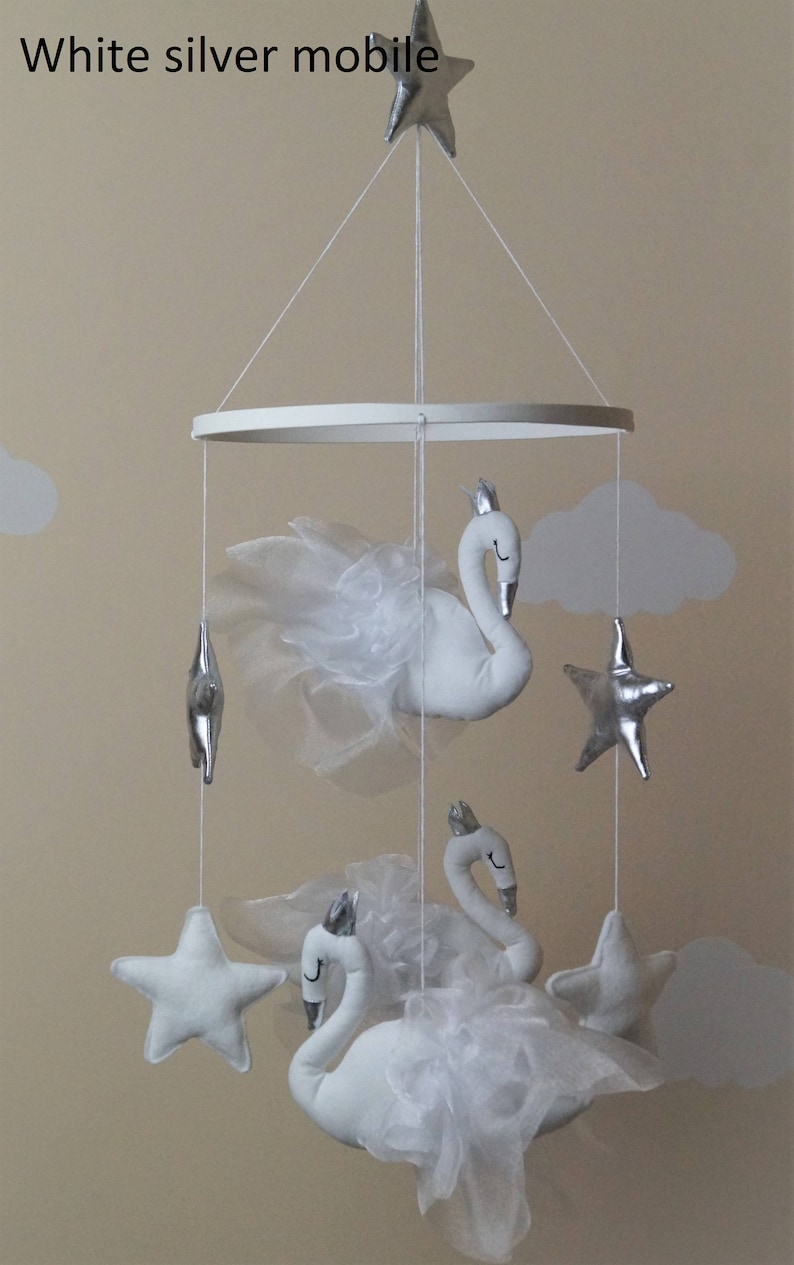 Puede incluir: M&oacute;vil de beb&eacute; blanco y plateado con tres cisnes blancos y tres estrellas plateadas. Los cisnes tienen alas de tul blanco y coronas. El m&oacute;vil est&aacute; colgado de un anillo blanco.