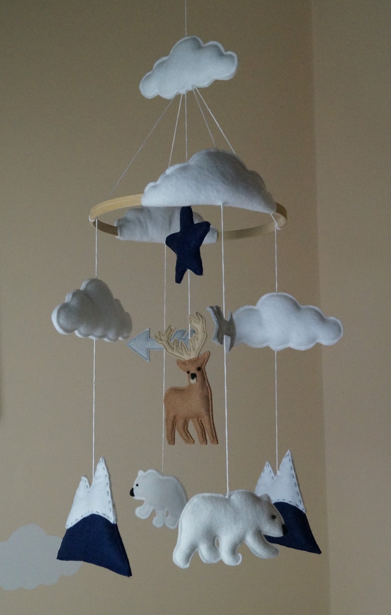 May include: A handmade baby mobile featuring felt clouds, a navy blue star, a brown deer, white polar bears, and navy blue and white mountains. The mobile is suspended from a wooden hoop with white string.