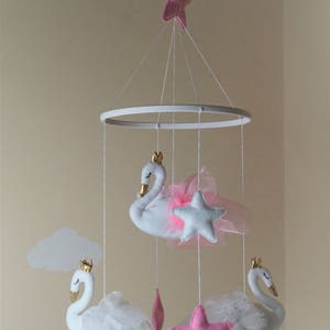 Puede incluir: Un m&oacute;vil para beb&eacute; blanco y rosa con tres cisnes blancos con coronas doradas y tul rosa, y tres estrellas rosas. El m&oacute;vil est&aacute; colgado de un techo blanco.