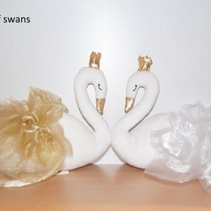 Puede incluir: Un par de figuras de cisnes blancos con coronas doradas, cada uno con una falda de tul. El cisne de la izquierda tiene una falda de tul dorada y el cisne de la derecha tiene una falda de tul blanca.