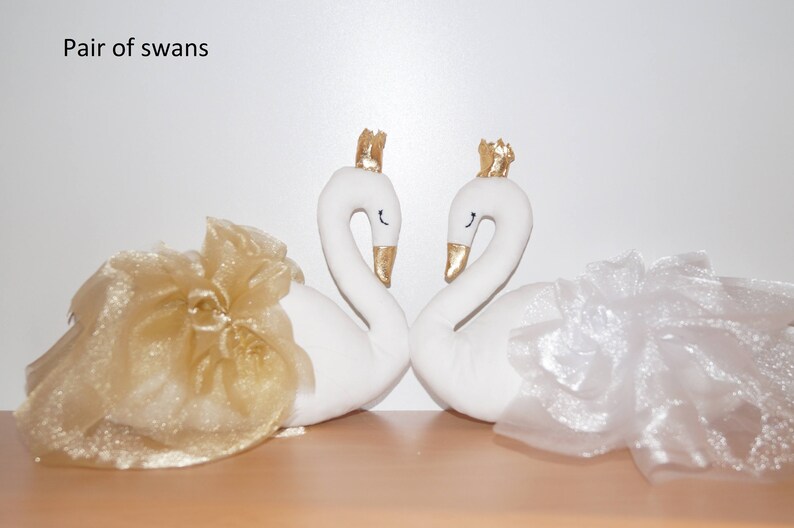 Puede incluir: Un par de figuras de cisnes blancos con coronas doradas, cada uno con una falda de tul. El cisne de la izquierda tiene una falda de tul dorada y el cisne de la derecha tiene una falda de tul blanca.
