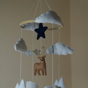 May include: A handmade baby mobile featuring felt clouds, a navy blue star, a brown deer, white polar bears, and mountains. The mobile is suspended from a wooden hoop with white string, creating a whimsical nursery decoration.