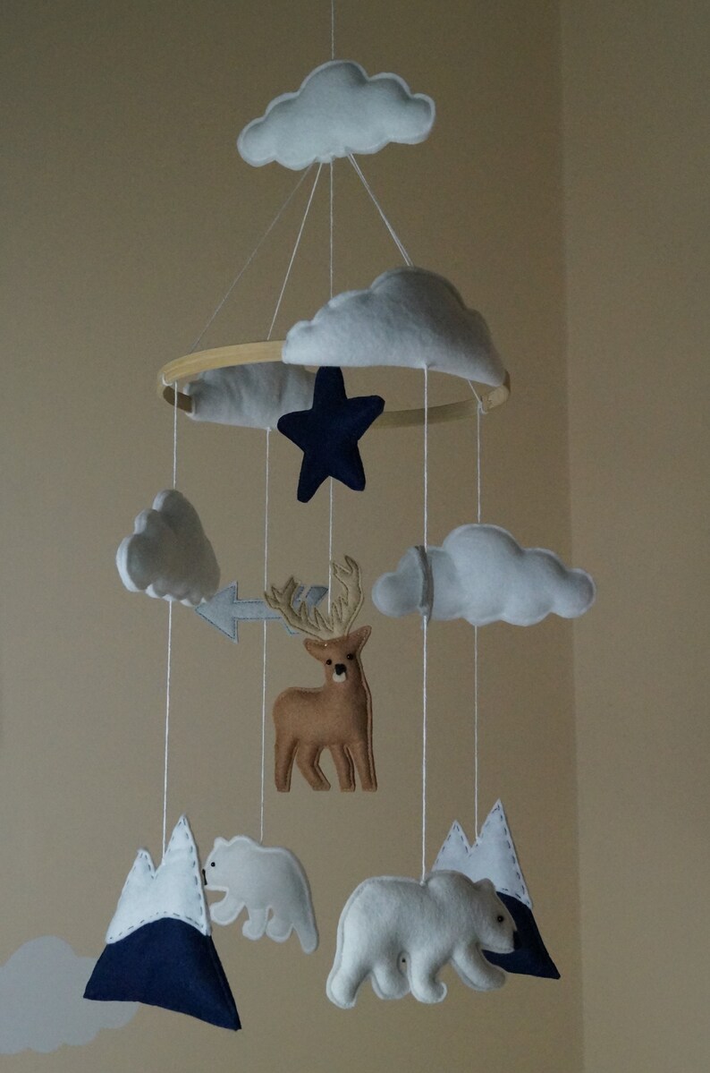 May include: A handmade baby mobile featuring felt clouds, a navy blue star, a brown deer, white polar bears, and mountains. The mobile is suspended from a wooden hoop with white string, creating a whimsical nursery decoration.