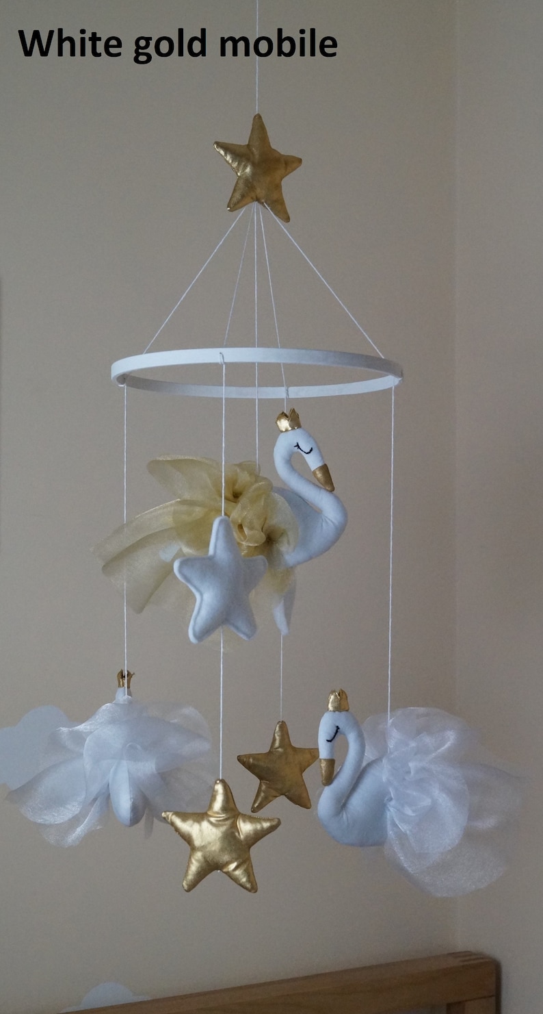 Puede incluir: M&oacute;vil de oro blanco con dos cisnes blancos, estrellas doradas y nubes blancas. El m&oacute;vil est&aacute; colgado de un anillo blanco.