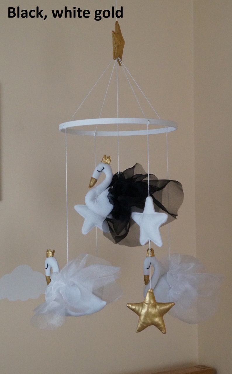 Puede incluir: Un m&oacute;vil para beb&eacute; blanco y dorado con tres cisnes blancos y tres estrellas doradas. Los cisnes tienen tul negro y blanco y coronas doradas.
