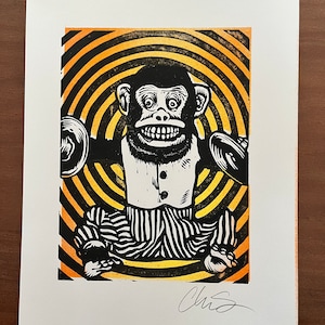 May include: A black and white linocut print of a monkey wearing a tuxedo and playing cymbals. The monkey is surrounded by concentric circles in black and gold.