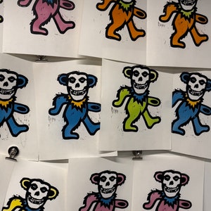 May include: Nine colorful prints of a cartoon bear with a skull face. The bear is walking and has a yellow, pink, blue, green, or orange collar. The prints are on white paper.