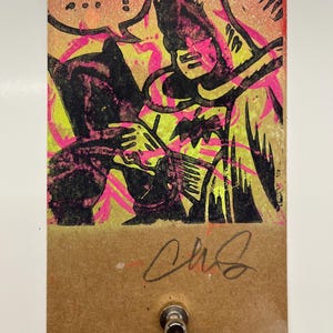May include: A colorful, abstract art print featuring a black figure with a bat symbol, set against a vibrant yellow and pink background. The artwork includes speech bubbles and a signature. A metal button is attached to the bottom.
