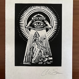 May include: Black and white linocut print featuring an eye within a triangle, radiating light onto a robed figure. The print has a detailed, graphic style with a signature in the lower right corner. The artwork is on white paper.