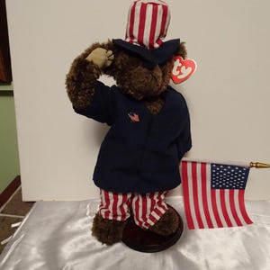 Puede incluir: Un oso de peluche marrón vestido como el Tío Sam, con una chaqueta azul marino con un pin de la bandera estadounidense, pantalones a rayas rojas y blancas y un sombrero de copa a juego. El oso sostiene una pequeña bandera estadounidense.