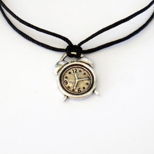 Könnte beinhalten: Ein schwarzes Armband aus Schnur mit einem Wecker-Anhänger. Das Zifferblatt der Uhr ist silberfarben mit schwarzen Zahlen und Zeigern.