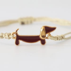 Könnte beinhalten: Ein Armband mit einem zentralen, goldfarbenen Metallanhänger in Form eines Dackels mit dunkelroter Emaillefüllung. Der Anhänger ist an einer dünnen, schimmernden goldfarbenen Kordel befestigt, die auf beiden Seiten verstellbare Schiebeknoten aufweist.